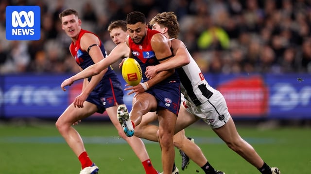 A Melbourne AFL player is tackled from the side by a Collingwood player as he tries to get a kick away.