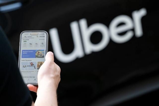 Ein Mann steht vor einem Mietwagen mit einem Schriftzug des Mobilitätsdienstleisters und Fahrtenvermittlers Uber und hält sein Smartphones mit der App des Unternehmens in der Hand.