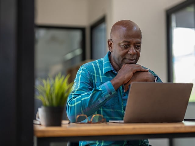 A man appears to be thinking while sitting at his desk in front of his laptop.