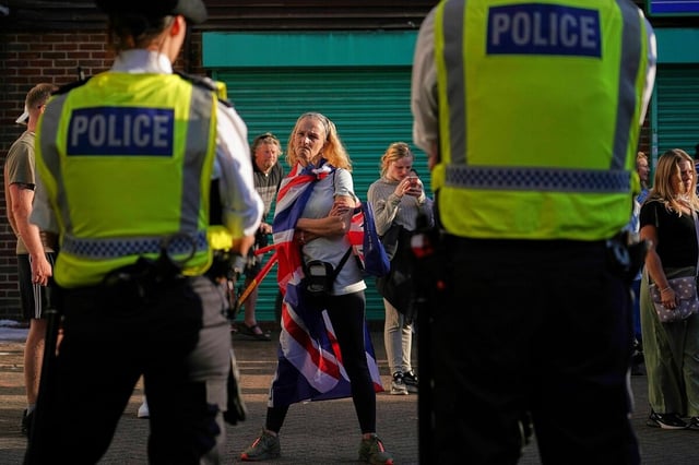 Eine Frau steht zwei Polizisten gegenüber, sie verschränkt ihre Arme und hat ein wütendes Gesicht. Über hren Schultern hängt die Fahne Großbritanniens