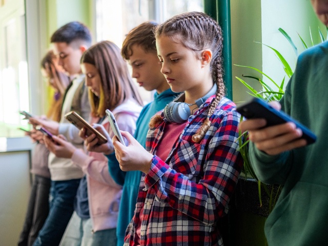 Teenage students are standing in the school hallway, all looking at their phones.