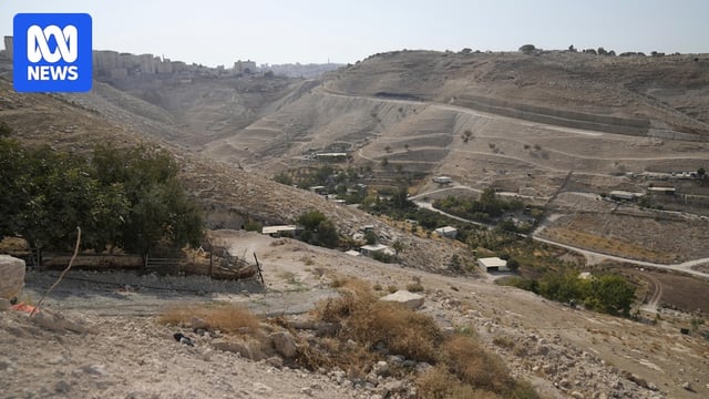 A dusty valley with small roads and paths cut into the side of a hillside, which sits beside a small collection of buildings.
