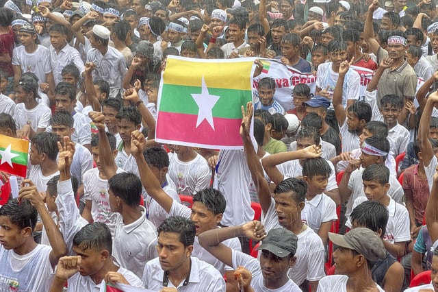 FILE - Rohingya refugees gather in the rain, with a flag of Myanmar, center, to demand safe return to Myanmar's Rakhine state as they mark the seventh anniversary of their mass exodus at their refugee camp at Kutupalong in Cox's Bazar district, Bangladesh, Aug. 25, 2024. (AP Photo/Shafiqur Rahman, File)