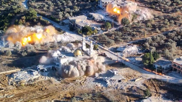 An aerial shot of a mosque and two other buildings exploding. Huge plumes of smoke and fire can be seen coming from the buildings. Two of the buildings are separated from other by a straight road which runs diagonally from the bottom right hand corner of the image. The buildings set among dusty earth and green foliage