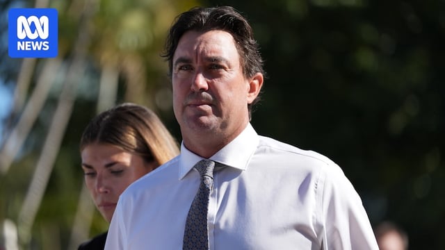 A close up shot of a white man, dark brown hair, tense expression, white button up shirt, tie. Blurred green background.