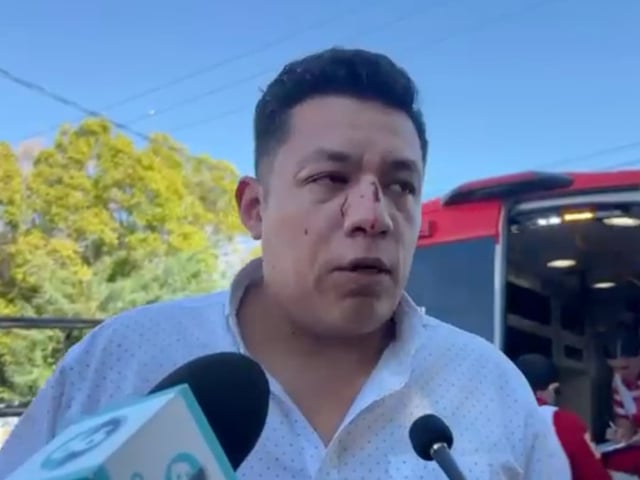 Jorge Heras, periodista de Mexicali, aparece con heridas visibles en el rostro mientras ofrece declaraciones a medios, luego de haber sido agredido por dos sujetos disfrazados de barrenderos. Al fondo se observa una ambulancia y personal médico atendiendo la situación.