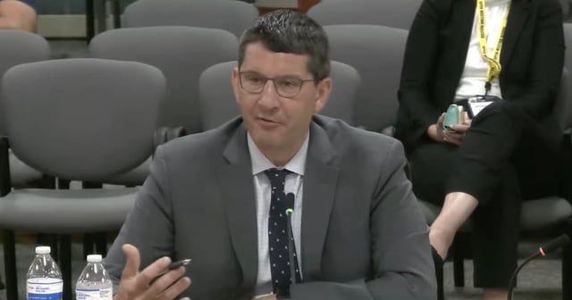 A screenshot of a white man in a grey suit sitting at a table speaking during an interview.