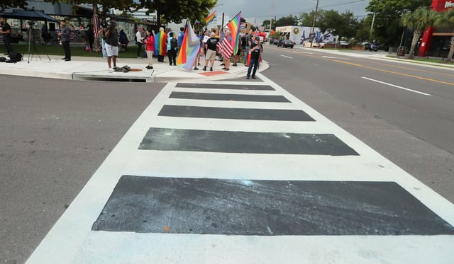 Demonstrators wave flags and cheer during a protest at a crosswalk in front of the former Pulse nightclub in Orlando on Thursday, August 21, 2025. The crosswalk had been painted in rainbow-themed colors, but overnight Wednesday, the rainbow-colored paint was removed from the crosswalk by the Florida Department of Transportation in an ongoing effort to remove “political banners” from public roadways. This sparked outrage from the LGBTQ+ community. During Thursday’s protest, demonstrators “replaced” the rainbow colors on the crosswalk using chalk. The former Pulse gay nightclub is now a memorial site for the 49 victims who were murdered there in 2016. (Stephen M. Dowell/Orlando Sentinel)