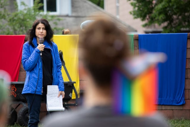 AG Dana Nessel is on a platform holding a mic and addressing a crowd of protesters. Around her are flags including the LGBT Rights flag.