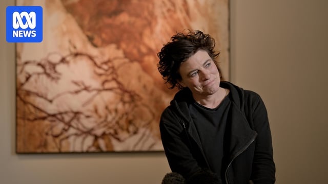 A woman in black clothing stands in front of a red dirt artwork.