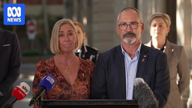A blonde woman in an orange top and a man with a beard in a suit speaking at a podium