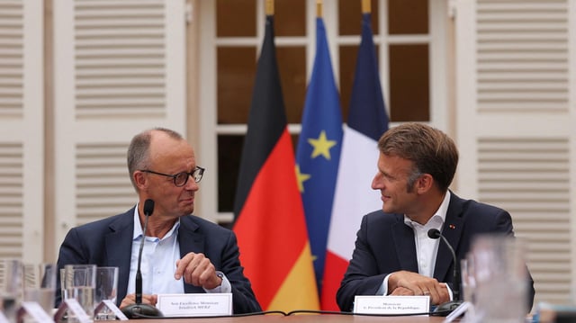 Friedrich Merz und Emmanuel Macron sitzen nebeneinander und schauen sich an.