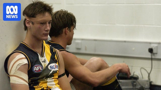 A downcast young AFL player with a bandage on his head leans against a wall next to a teammate.