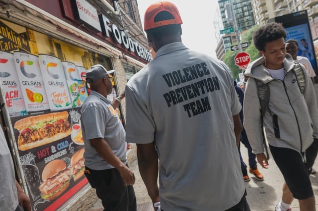 A man stands on the sidewalk in Crown Heights with his back to the camera wearing a gray shirt that says VIOLENCE PREVENTION TEAM.