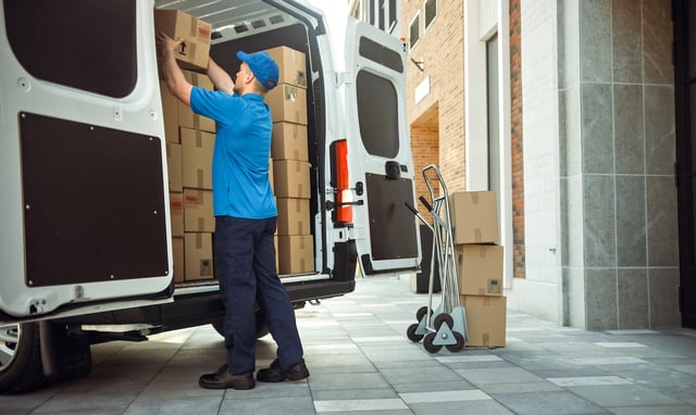 Delivery Man Uses Hand Truck Trolley Full of Cardboard Boxes and Packages, Loads Parcels into Truck / Van. Professional Courier / Loader helping you Move, Delivering Your Purchased Items Efficiently