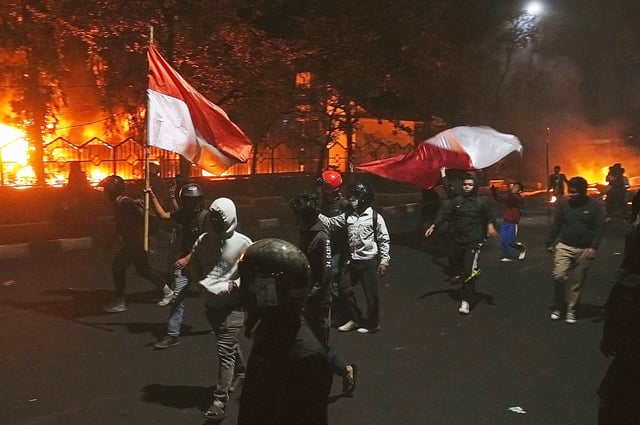 Protesters walk by as the local parliament building is engulfed in flames during a protest following the death of a delivery rider in clashes between riot police and students protesting against lawmakers' allowances in Makassar, South Sulawesi, Indonesia, Friday, Aug. 29, 2025. (AP Photo/ Masyudi Firmansyah)