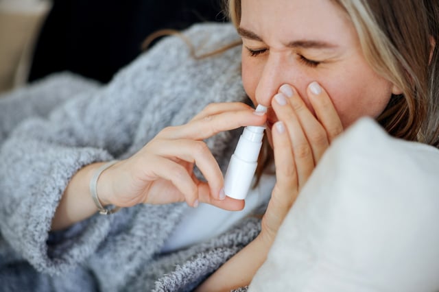 Caucasian woman is shown using a nasal spray in her bedroom to alleviate her congestion.