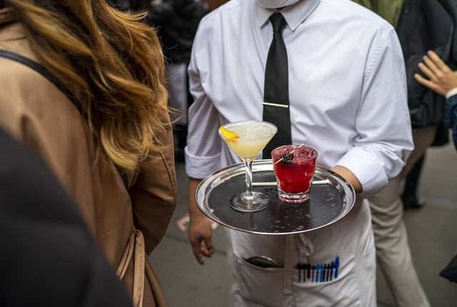 A waiter wearing a mask carries drinks across a sidewalk to an outdoor table at Balthazar Restaurant November 13, 2021 in New York City.