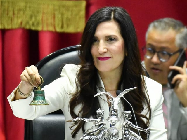 Kenia López Rabadán sostiene la campana presidencial tras asumir como presidenta de la Mesa Directiva de la Cámara de Diputados, durante una sesión oficial en el recinto legislativo. Viste de blanco y se le ve sonriente mientras preside la sesión, con fondo rojo institucional y ornamentos legislativos visibles.