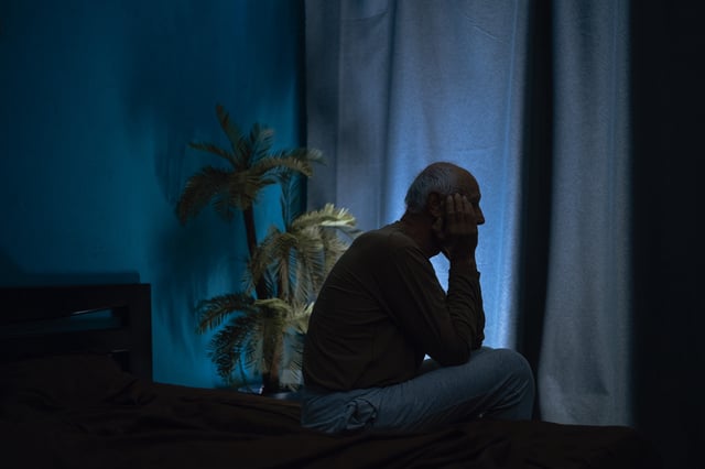 A stock image of a silhouette of an old man suffering from insomnia sitting on his bed at night.