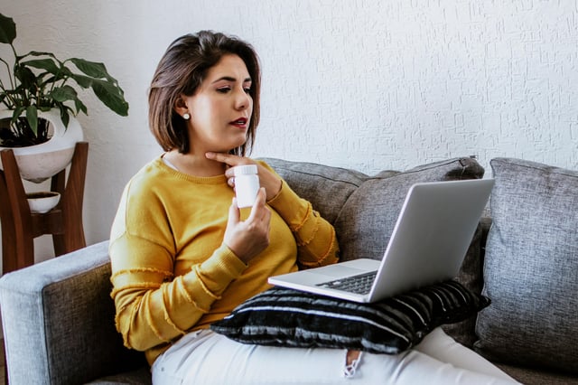 Woman sitting on sofa using laptop to video call doctor while holding pills.