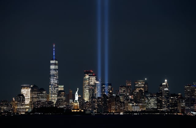 The New York City skyline at night with the Tribute in Light memorial shining two bright blue beams into the sky, the Statue of Liberty illuminated in the foreground, and One World Trade Center lit up to the left.