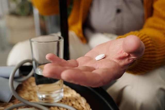 Man holding pill in open palm, sitting at table with glass of water and blood pressure monitor.
