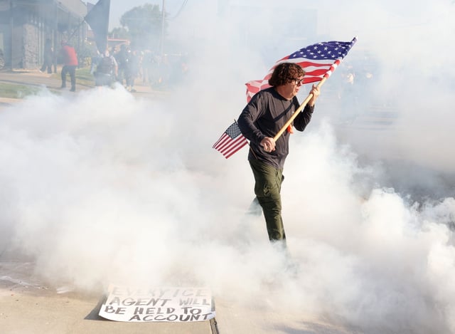 Curtis Evans, of Evanston, a military veteran USMC intelligence, during President Reagan’s era, carries a U.S. Flag through deterrents deployed by federal officers as they clear protesters from the entrance of the Immigration and Customs Enforcement facility in Broadview, Sept. 19, 2025.  (Stacey Wescott/Chicago Tribune)