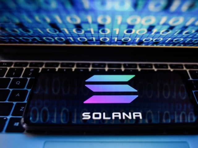 In this photo illustration a Solana logo seen displayed on a smartphone with binary code on a laptop screen