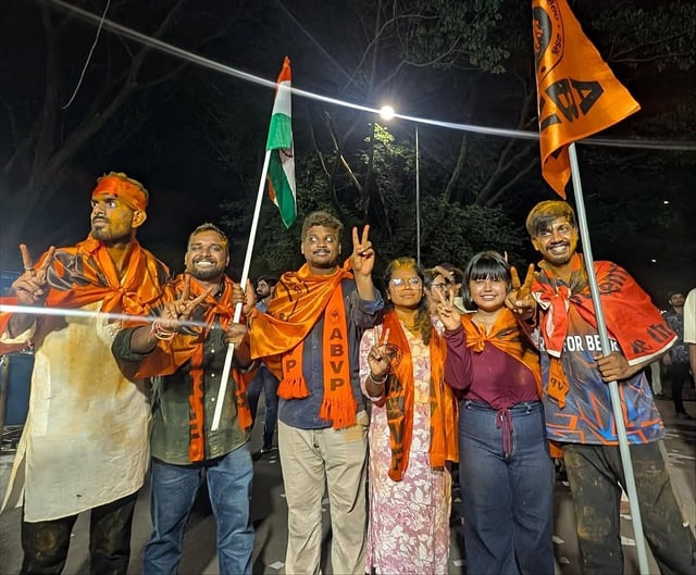 A group of six young people standing together at night, smiling and making peace signs with their hands. They are wearing bright orange scarves and shawls with “ABVP” written on them, and some have colored powder smeared on their faces and clothes. One person holds the Indian national flag, while another holds an orange ABVP flag. The background shows trees and streetlights, suggesting the photo was taken outdoors during a celebration or gathering.