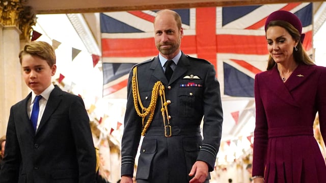 Prince George wears a suit, Prince William wears military uniform, Kate Middleton wears a dark red coat dress, as they walk in front of a Union Jack flag