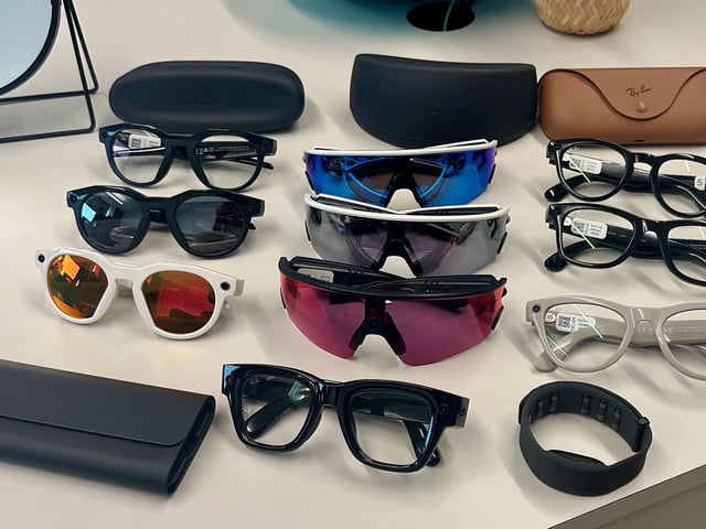 Three Oakley Meta HSTN, three Oakley Meta Vanguard, and three Ray-Ban Meta (Gen 2), along with cases, sitting atop a table in various styles, along with the Meta Ray-Ban Display glasses in front of them all.