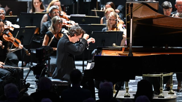 A pianist in black performs on stage with an orchestra. He leans forward, eyes closed, playing a grand piano. Musicians with violins and cellos are visible behind him. Audience members sit in the foreground.
