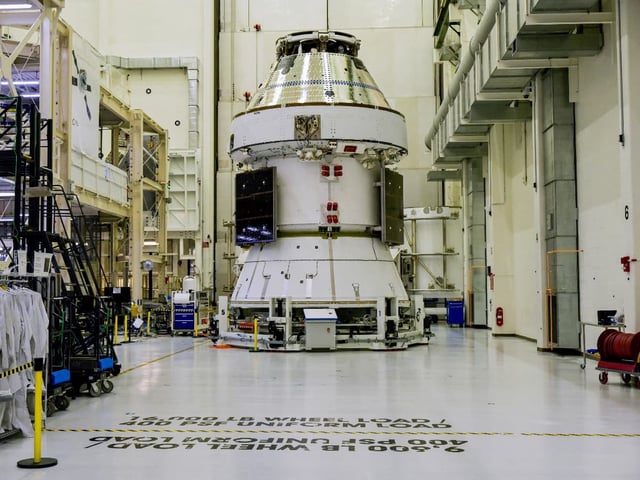 El módulo tripulado Orión de la misión Artemis 2 de la NASA, dentro del edificio de operaciones del Centro Espacial Kennedy, rodeado de infraestructura técnica y trajes de astronauta, durante un evento de medios previo al lanzamiento previsto para 2026
