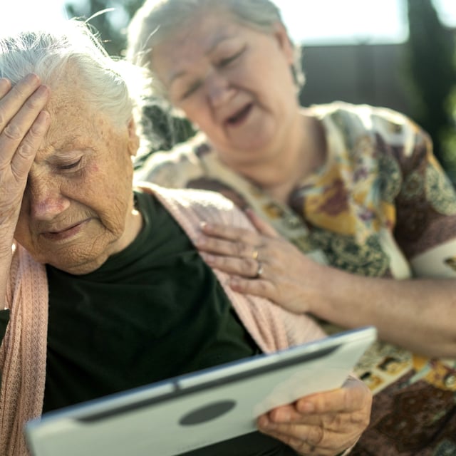 An elderly sad woman in a wheelchair looks at a tablet and talks to her family via video call - stock photo
