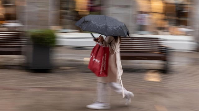 Eine Frau mit Schirm geht durch eine nasse Einkaufsstraße. 