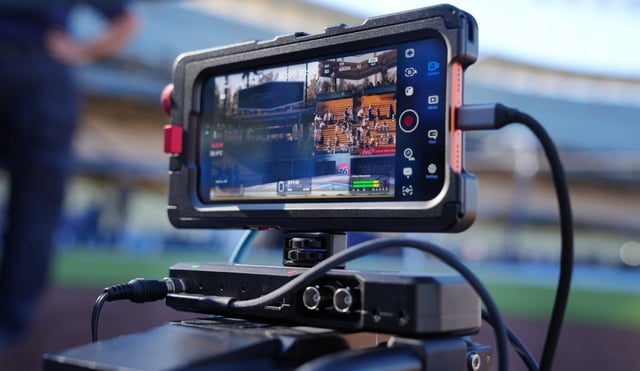 A smartphone mounted on a camera rig displays a live sports event. The screen shows a stadium with spectators and a field. The rig includes cables and a battery pack for power.