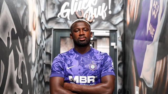 Ein Fußballer steht in einem Gang mit verschränkten Armen. Im Hintergrund steht an der Wand über einer Tür: Glück auf.