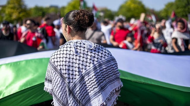 Eine junge Frau mit Kufiya-Tuch um die Schultern hilft eine große Palästinaflagge zu tragen, im Hintergrund sind weitere Demonstranten zu sehen