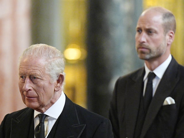 Prince William and King Charles wearing black suits looking somber