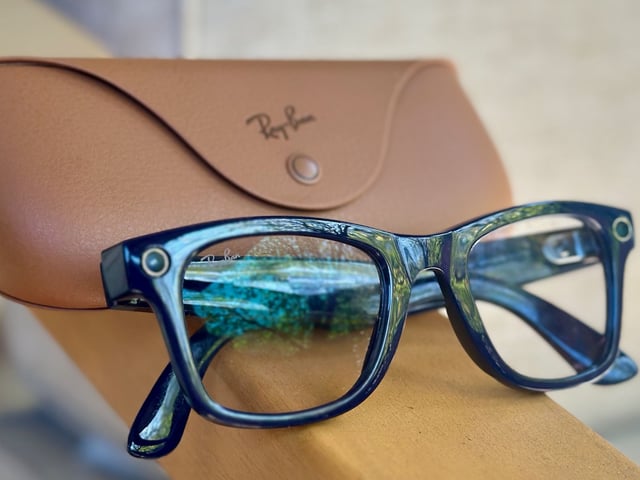 Close-up photo of the Shiny Cosmic Blue Ray-Ban Meta (Gen 2) Wayfarer glasses sitting folded up on an armrest with the official brown Ray-Ban charging case behind it.