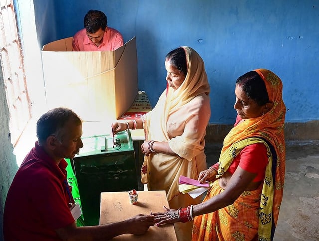 A man in a red polo shirt is seated at a table, interacting with two women during an election. The man is holding the hand of one of the women, who is wearing a bright orange and yellow sari and holding a small stack of papers or ballots. The second woman, wearing a cream-colored headscarf and shawl, is standing between the man and the other woman, and appears to be interacting with a small electronic device or ballot box on the table. In the background, another man in a pink shirt is partially visible behind a cardboard voting screen. A sign in Hindi is posted on the light blue wall on the left. The scene is brightly lit by a window on the left.