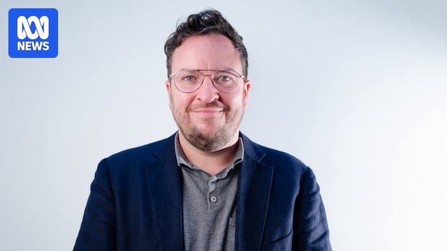 Rick Morton, a man in his late 30s with stubble, smiles slightly, wearing a blazer and wire-framed glasses.