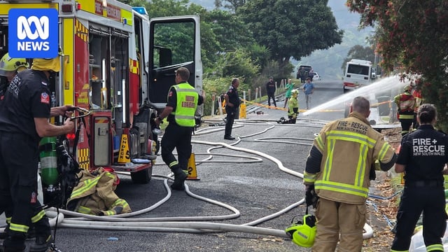 Uniformed firefighters move around a fire truck while one firefighter kneels on the ground pointing a fire hose off camera.