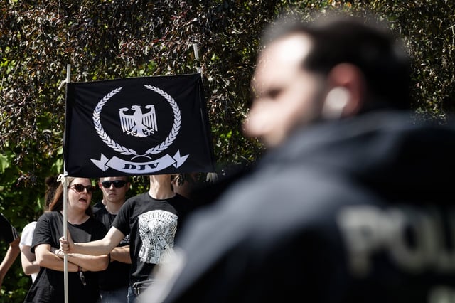 DJV-Aufmarsch in Berlin: Ein Polizist im Vordergrund, hinten halten Menschen eine schwarze Fahne mit Adler und Aufdruck DJV