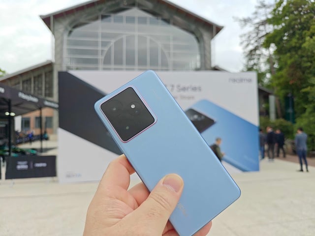 Hand holding blue Realme GT 7 with phone poster in background