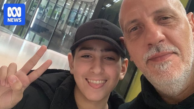 A young boy in a black had gives the peace sign in a close-up selfie with his dad, a man with grey facial hair.  