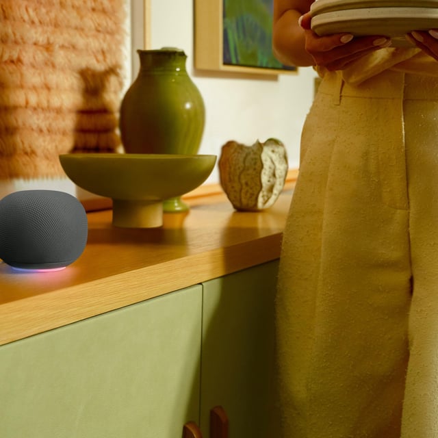 The Google Home Speaker in black, sitting on a countertop while a model walks by.