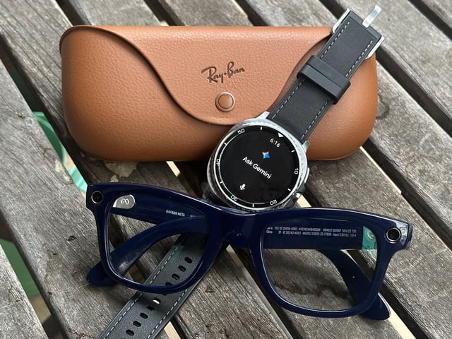 A photo of the Ray-Ban Meta (Gen 2) smart glasses and case sitting on a table with the Samsung Galaxy Watch 8 Classic between them, leaned against the case, with the display showing "Ask Gemini."