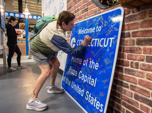 Connecticut has been touting its pizza on a constant basis. Here, Cheryl Sanderson of Columbia signs the "Welcome to Connecticut : Home of the Pizza Capital of the United States" sign inside the Connecticut building during Connecticut Day at The Big E on Sept. 17, 2025. (Aaron Flaum/Hartford Courant)